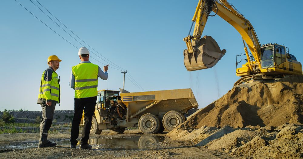 Male Real Estate Investor And Project Manager Talking On Construction Site Of Apartment Block. Colleagues Discussing Building Progress, Business. Excavator Loading Sand In Industrial Truck.