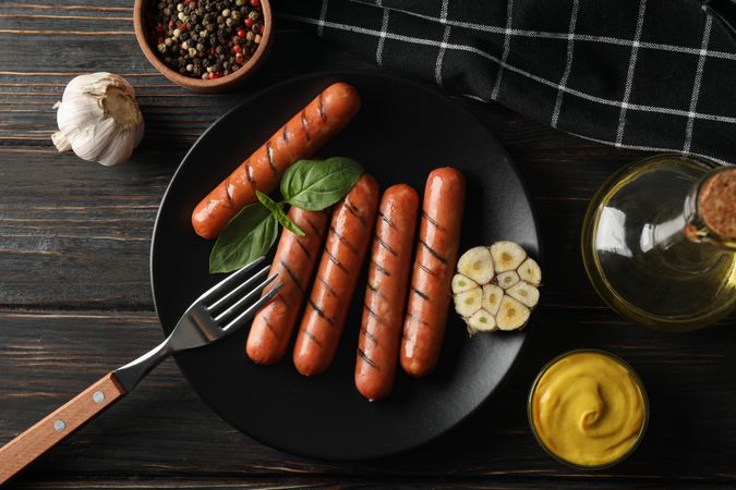 Top view of grilled sausages or hot dogs on a fork on dark ceramic plate with garlic