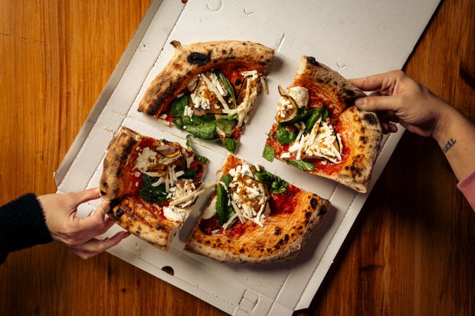 Four hands take pizza slices with cheese, spinach, and tomato sauce from a box