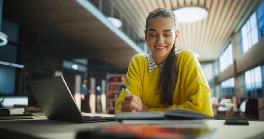 Happy and Focused Student Studying in a Modern Library. Young Female Enjoying Time Alone for Thinking and Problem Solving. Girl Using Laptop Computer to Work on a University Research Project Online