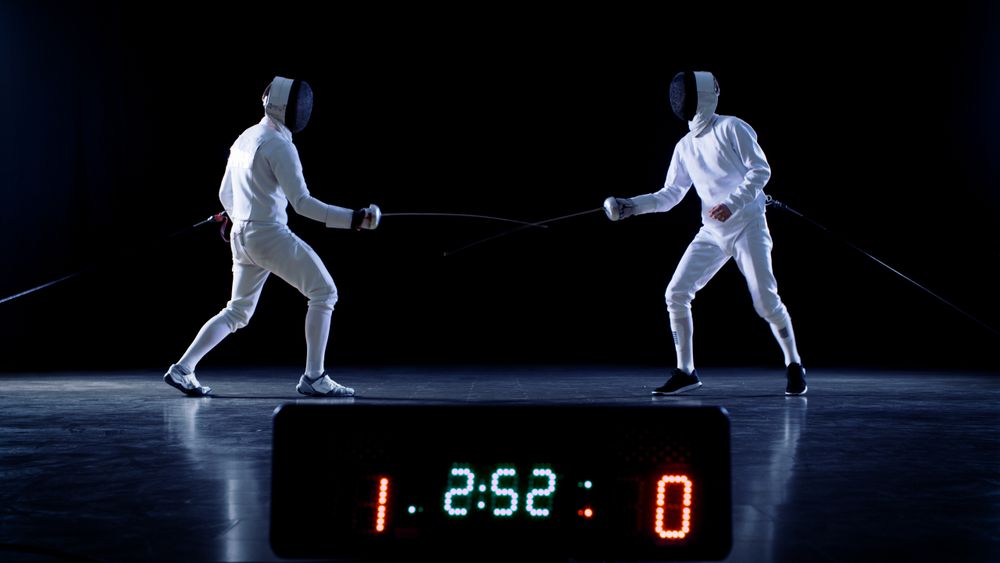 On the Championship Two Professional Fully Equipped Fencers Fight with Foils while Scoreboard Keeps the Scores of Hits.