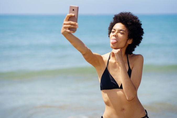 Woman in bikini taking silly selfie on phone on coast