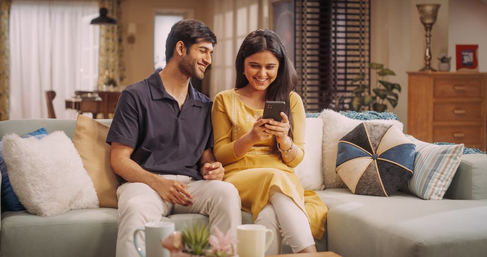 Young Indian Couple Using Internet On Smartphone, Sitting On The Sofa at Home, they Tease and Joke around.