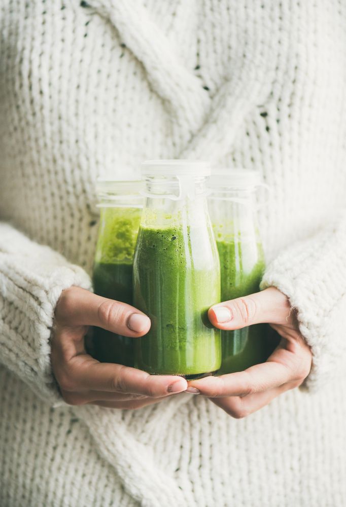 Woman holding three bottles of green smoothie wearing cozy sweater