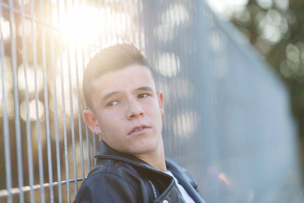 Teenage male wearing a leather jacket leaning on fence outside looking to the side