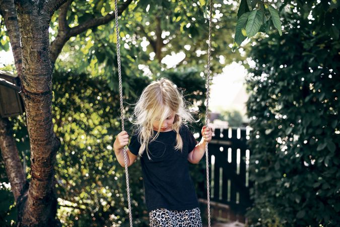 Shy blonde girl with braided hair standing on outdoor swing in backyard
