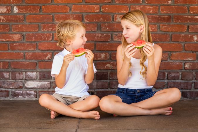 Cute Young Boy and Girl Eating Watermelon Against Brick Wall