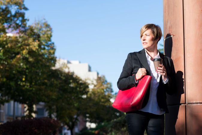 Female in blazer standing outside on sunny day with coffee and large red handbag