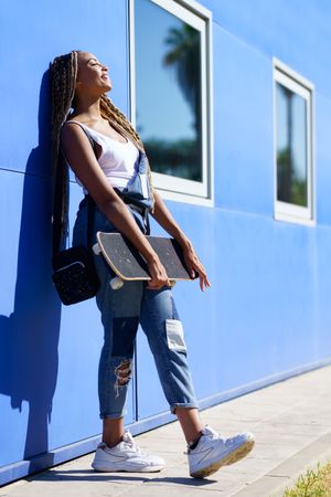 Female in denim overalls standing with skateboard while leaning on blue building, vertical