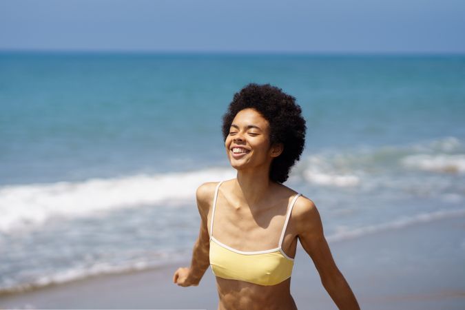 Woman walking on coast in yellow bikini with eyes closed to the sun