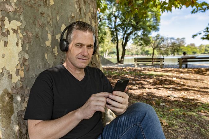 Man listening to music, sitting on the grass, and leaning on a tree