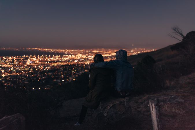 Couple watching the brightly lit city sitting on top of a hill late in the evening