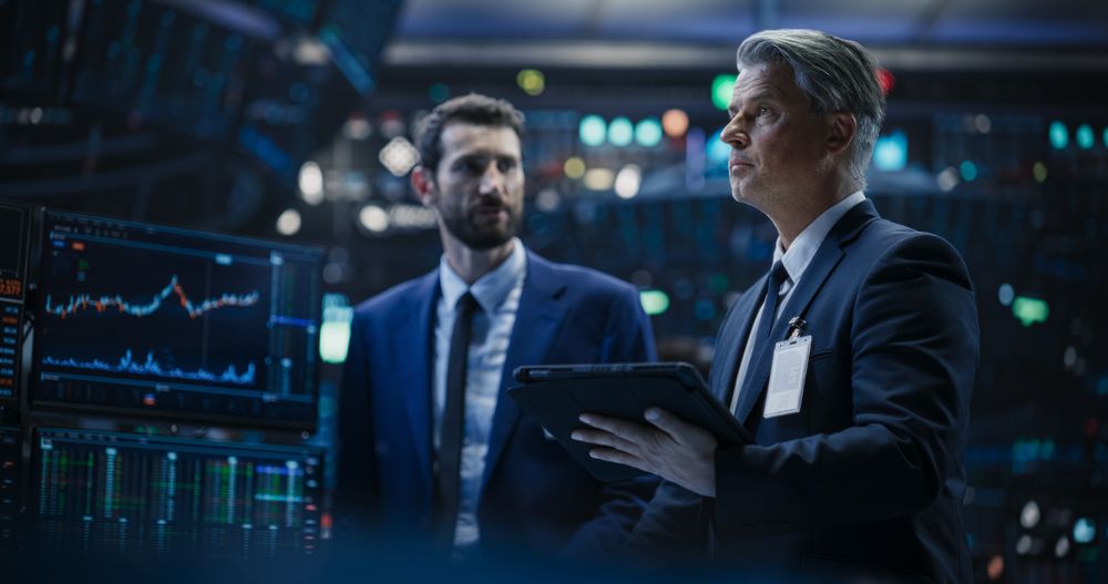 Team of Two Handsome Stock Exchange Brokers Using Tablet Computer, Chatting About the Asset Management Strategy for an International Business Client, Working in a Office Building with Computer Screens