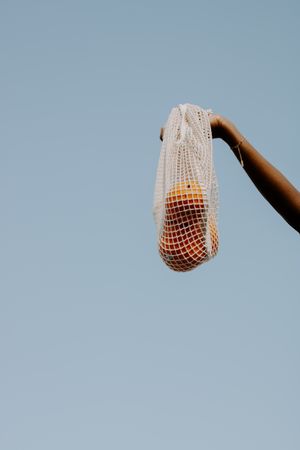 Person holding a mesh bag with oranges against blue background