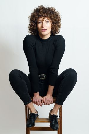 Woman dressed head to toe in dark color sitting on stool looking at camera