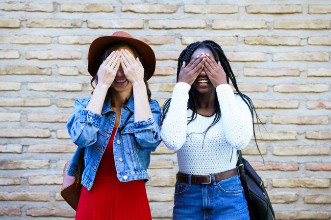 Two women covering their eyes before a brick wall