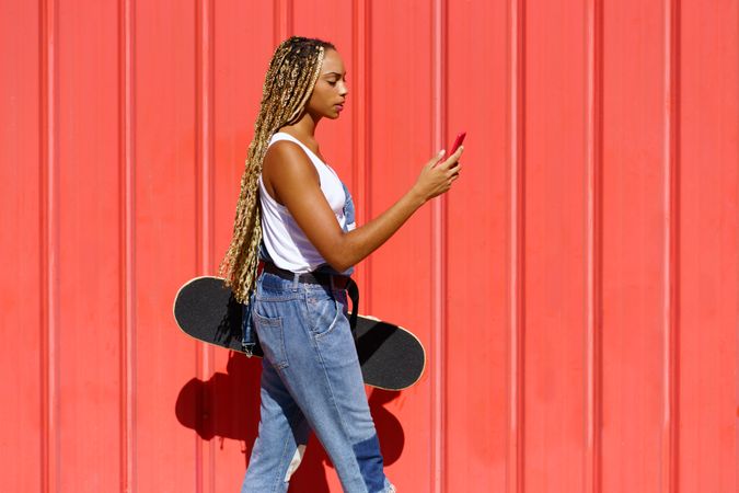 Woman walking in front of red wall texting on phone and holding a skateboard