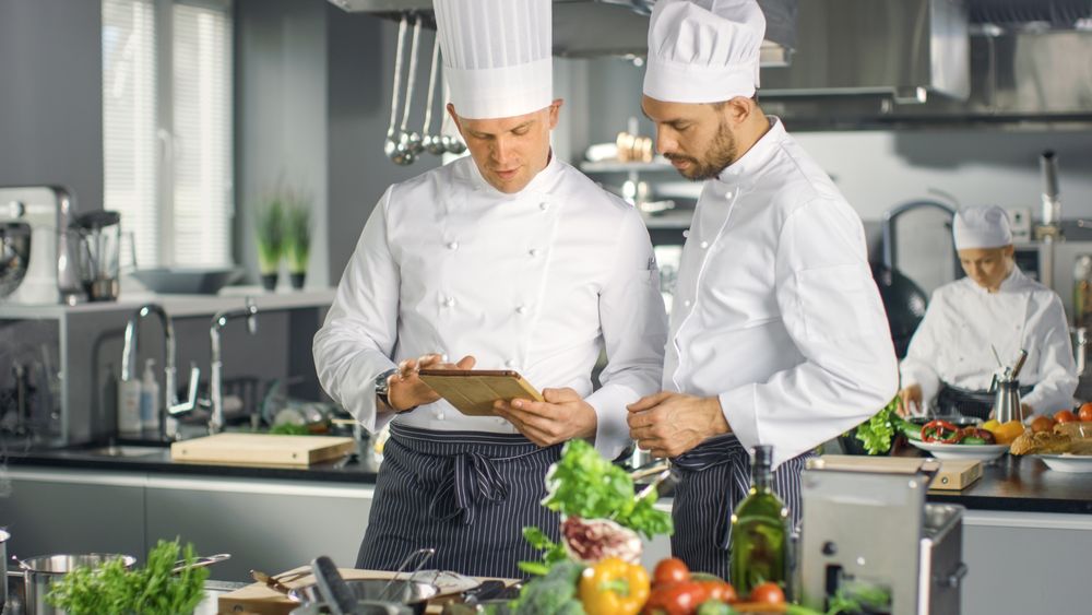 Two Famous Chefs Exchange Ideas About Video Blog Recipe they Watching on Tablet. They are Working in Big Restaurant Kitchen.