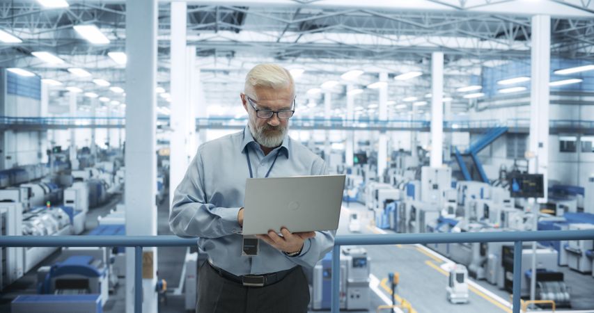 Factory Manager Working on Laptop at a Futuristic Industrial Manufacture with Autonomous AI Robots. Specialist Monitoring All the Data on a Computer, Checking Reports and Planning Workload