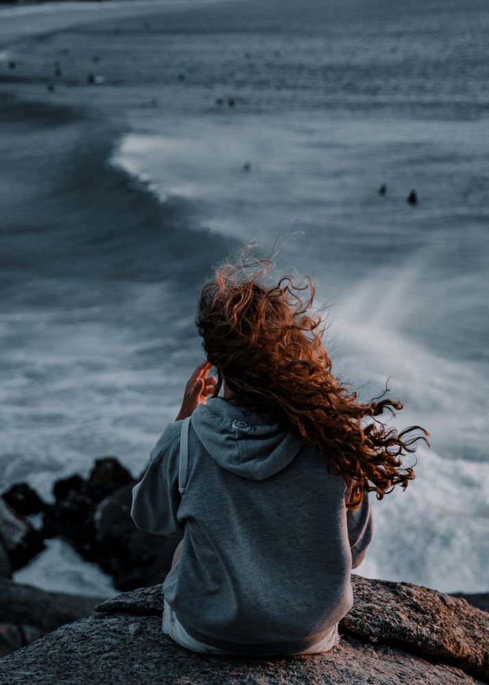 Back view of woman in gray hoodie sitting on rocky seashore