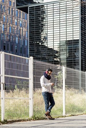 Male in headphones holding smartphone while leaning on a metallic fence outside
