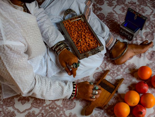 Man in traditional outfit sitting on floor beside orange fruit and tray of almond