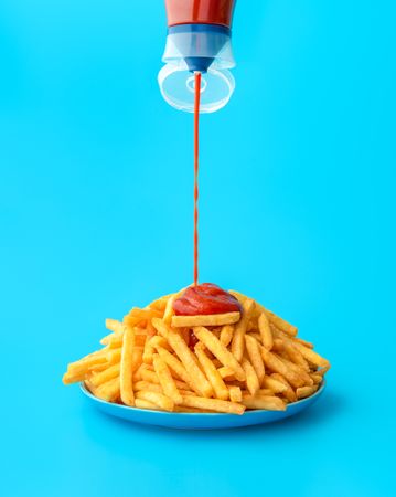 Pouring ketchup sauce over french fries, isolated on a blue background