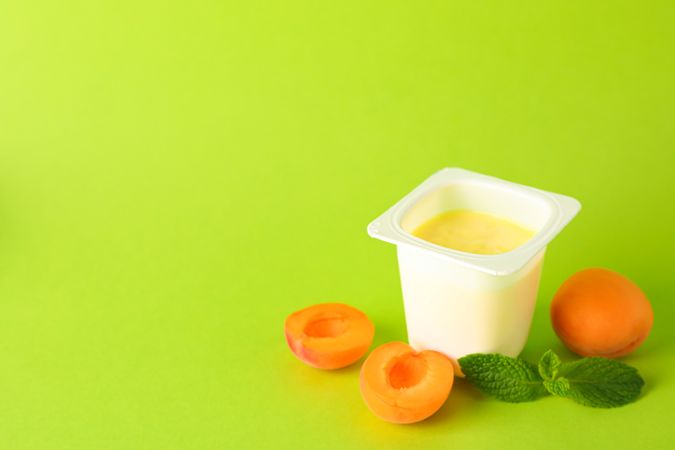 Plastic cup with yogurt and fruits on color background, space for text