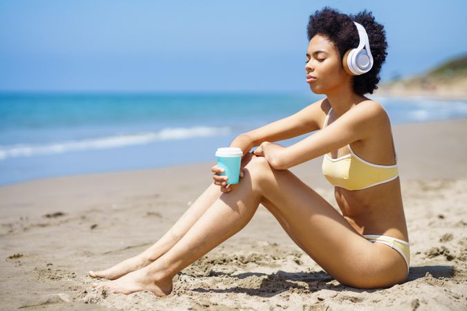 Calm woman in yellow bikini and headphones sitting on beach with coffee cup
