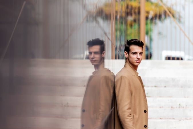 Side view of young man leaning on reflective wall outside looking at camera with space for text