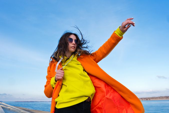 Trendy woman in sunglasses, orange coat, yellow hoodie and jeans in front of the ocean
