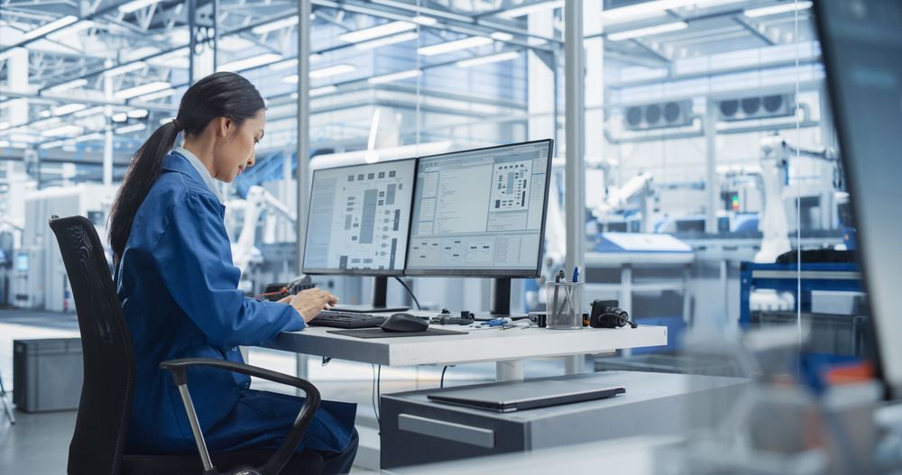 Experienced Female Software Developer Working in an Office, Using Desktop Computer and Overlooking Production at a Modern Automated Electronics Manufacture with Robotic Arms. Administrator at Work