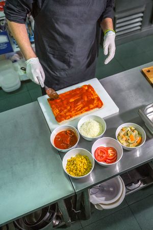 Anonymous male chef with black apron spreading tomato sauce over focaccia in restaurant kitchen