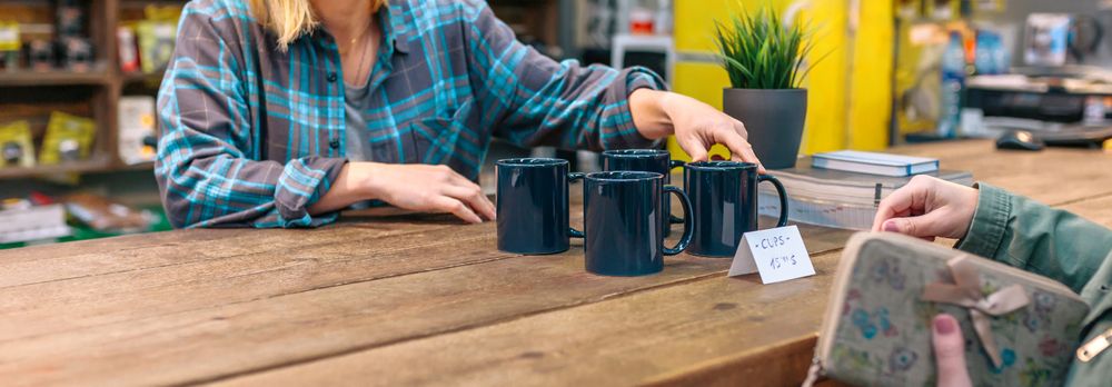 Banner of unrecognizable woman employee showing blue mugs to customer in vintage shop