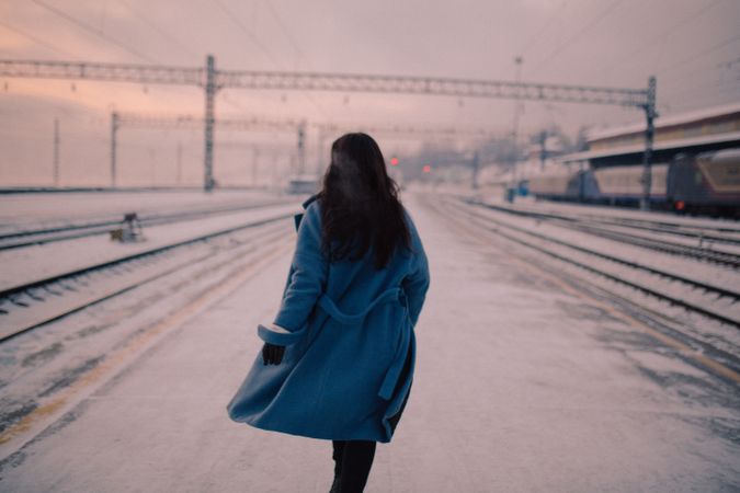 Back view of woman in blue coat walking on snow-covered ground at sunset