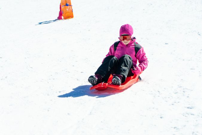 Child in pink snow suit going down a snowy hill in a plastic sled