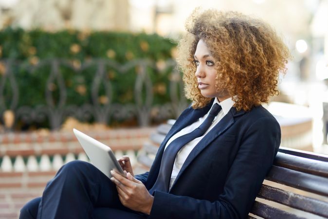 Woman in suit looking up from her digital tablet’s screen on outdoor bench