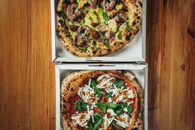 Two gourmet pizzas in cardboard boxes on a wooden table, topped with fresh ingredients