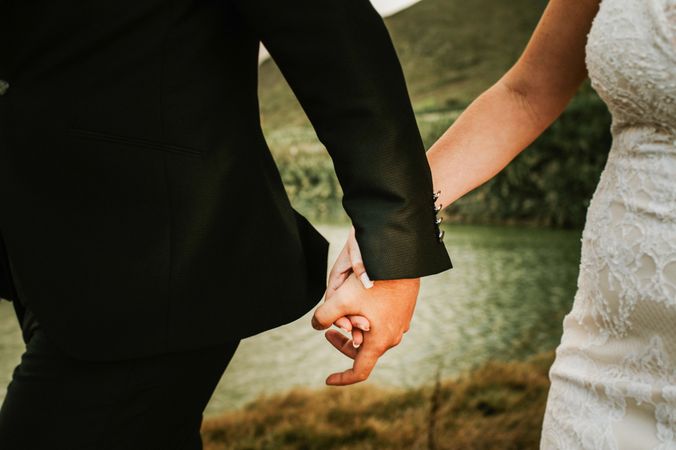 Crop groom and bride holding hands on seashore