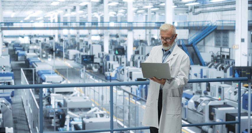 Experienced Industrial Scientist Wearing a White Coat, Using Laptop Computer and Overlooking Production at a Modern Automated Electronics Manufacture with Hydraulic Robotic Arms