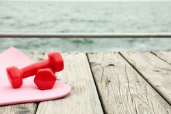 Yoga mat and dumbbells on wooden floor at sea, space for text