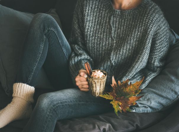 Woman in cozy sweater, socks and jeans holding whipped cream topped drink with fall leaves