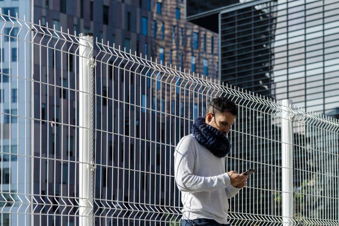 Young man wearing scarf leaning on metallic fence and checking smartphone outdoors