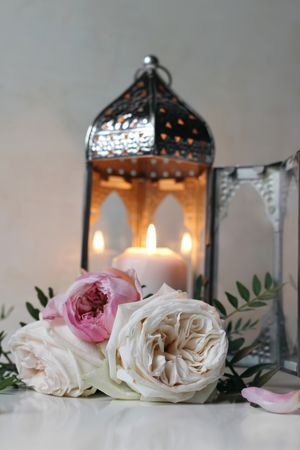Silver Moroccan, Arabic lantern with burning candle. Pink roses flowers on table. Festive still life for muslim holiday Ramadan Kareem or boho wedding. Blurred background, vertical.