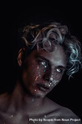Portrait of topless blonde man with UV paint on his face looking away against dark background 489wRb