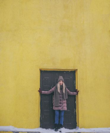 Woman in knit cap and coat standing beside door of yellow building