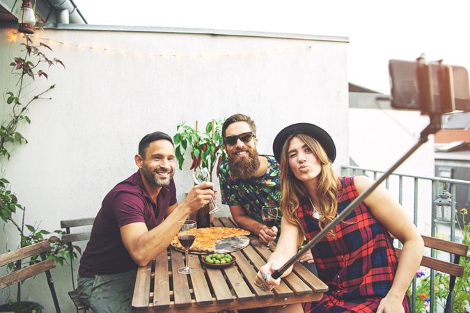 Group of friends enjoying pizza and wine on a rooftop patio with selfie stick