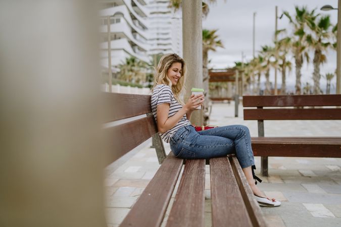 Happy woman with disposable cup of coffee sitting on street bench