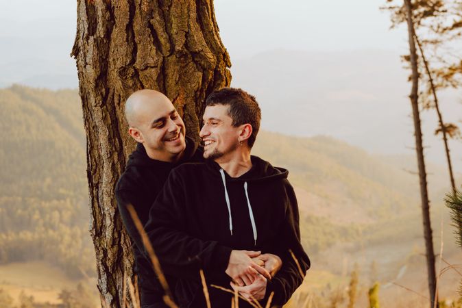 Smiling men hugging near tree in forest on hill