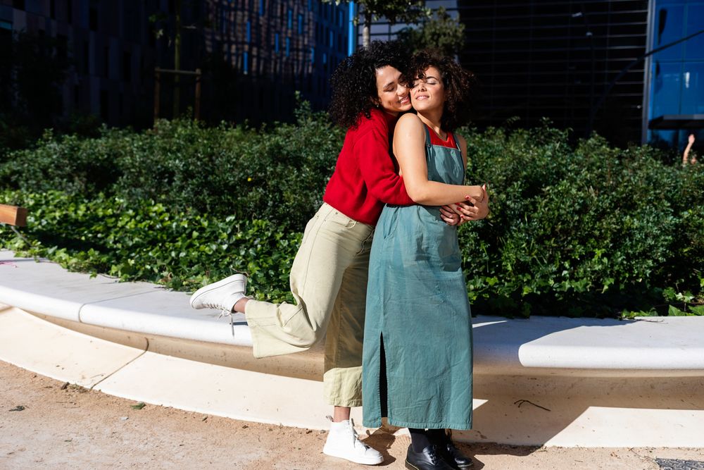 Fun female couple holding each other in city park with office buildings in background